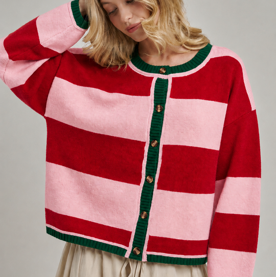 A woman wearing a pink and red striped knit cardigan with dark green trim and buttons, paired with a beige skirt, against a neutral gray studio background.