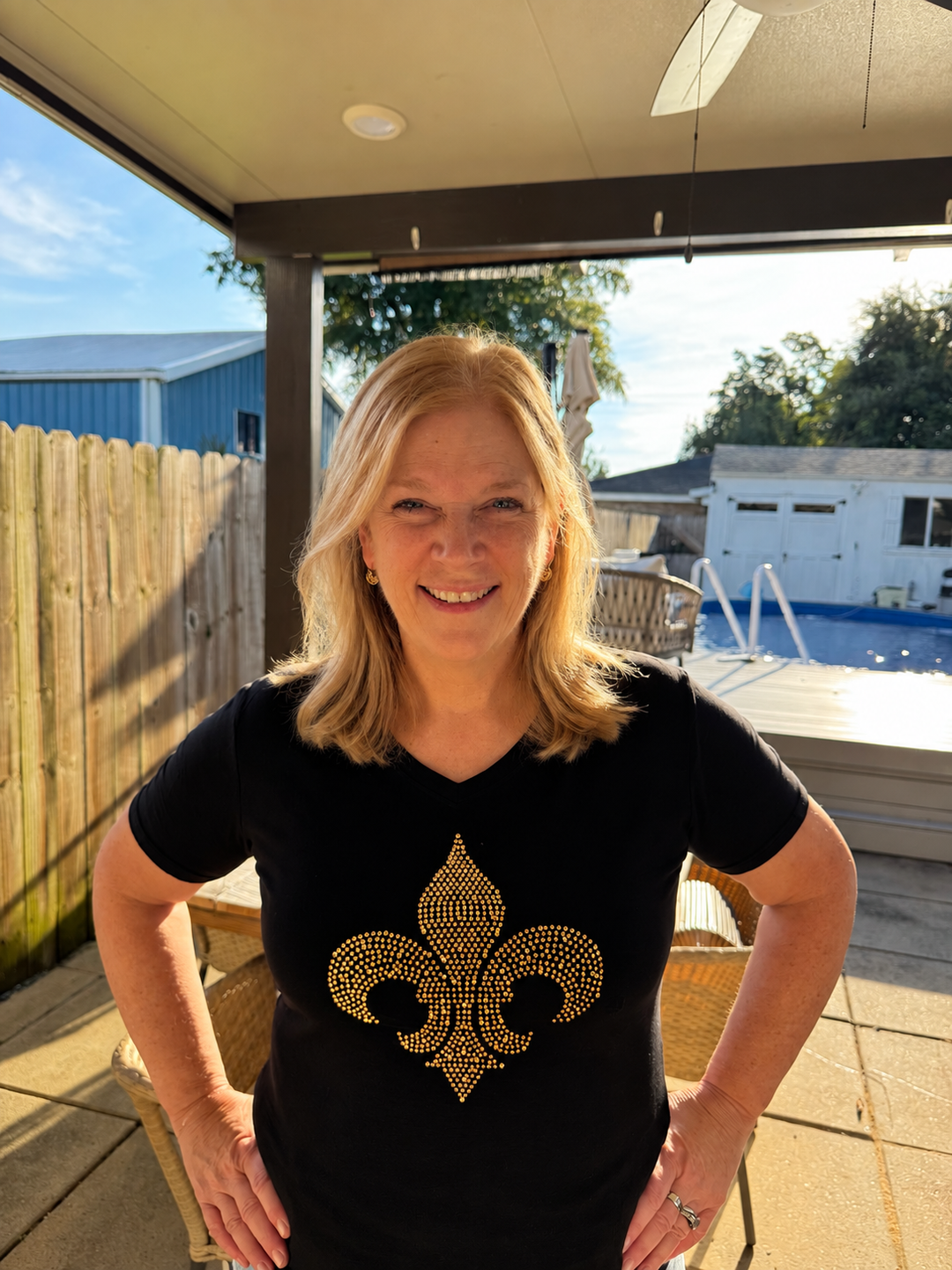 A woman wearing a black short-sleeve v-neck t-shirt with a large gold rhinestone fleur de lis graphic centered on the chest.