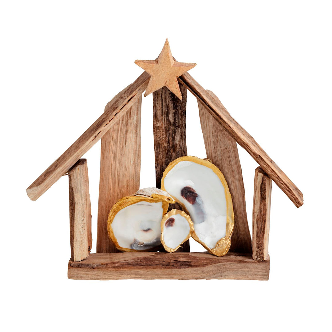 A rustic open-frame driftwood nativity stable topped with a carved wooden star, with three white oyster shells edged in gold arranged inside as the Holy Family figures.