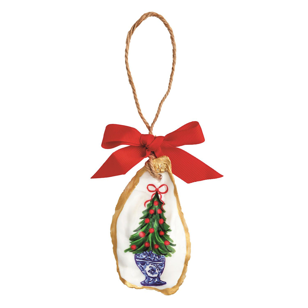 A hand-painted oyster shell Christmas ornament with a gold-painted edge, featuring a green Christmas tree topiary in a blue-and-white chinoiserie-style planter with red berries and a red bow, hung on a jute twine loop with a red grosgrain ribbon bow.