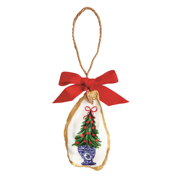 A hand-painted oyster shell Christmas ornament with a gold-painted edge, featuring a green Christmas tree topiary in a blue-and-white chinoiserie-style planter with red berries and a red bow, hung on a jute twine loop with a red grosgrain ribbon bow.