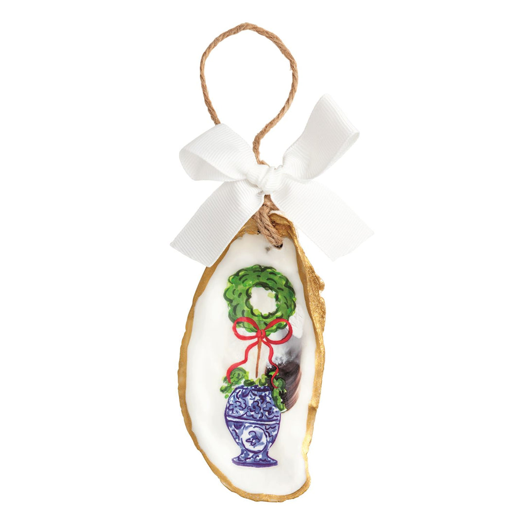 A hand-painted oyster shell Christmas ornament with a gold-painted edge, featuring a green Christmas tree topiary in a blue-and-white chinoiserie-style planter with red berries and a red bow, hung on a jute twine loop with a red grosgrain ribbon bow.