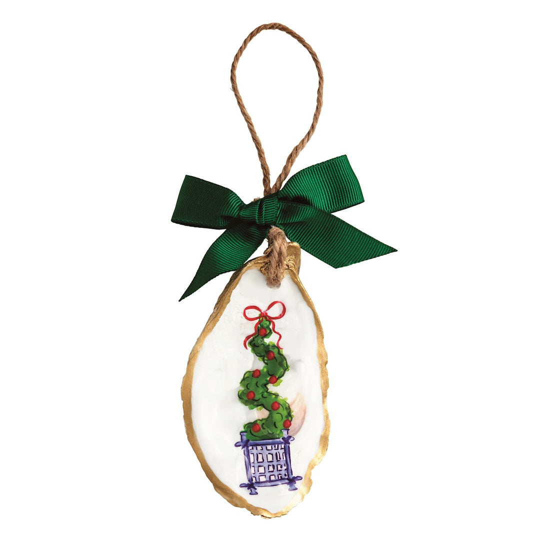 A hand-painted oyster shell Christmas ornament with a gold-painted edge, featuring a green Christmas tree topiary in a blue-and-white chinoiserie-style planter with red berries and a red bow, hung on a jute twine loop with a red grosgrain ribbon bow.