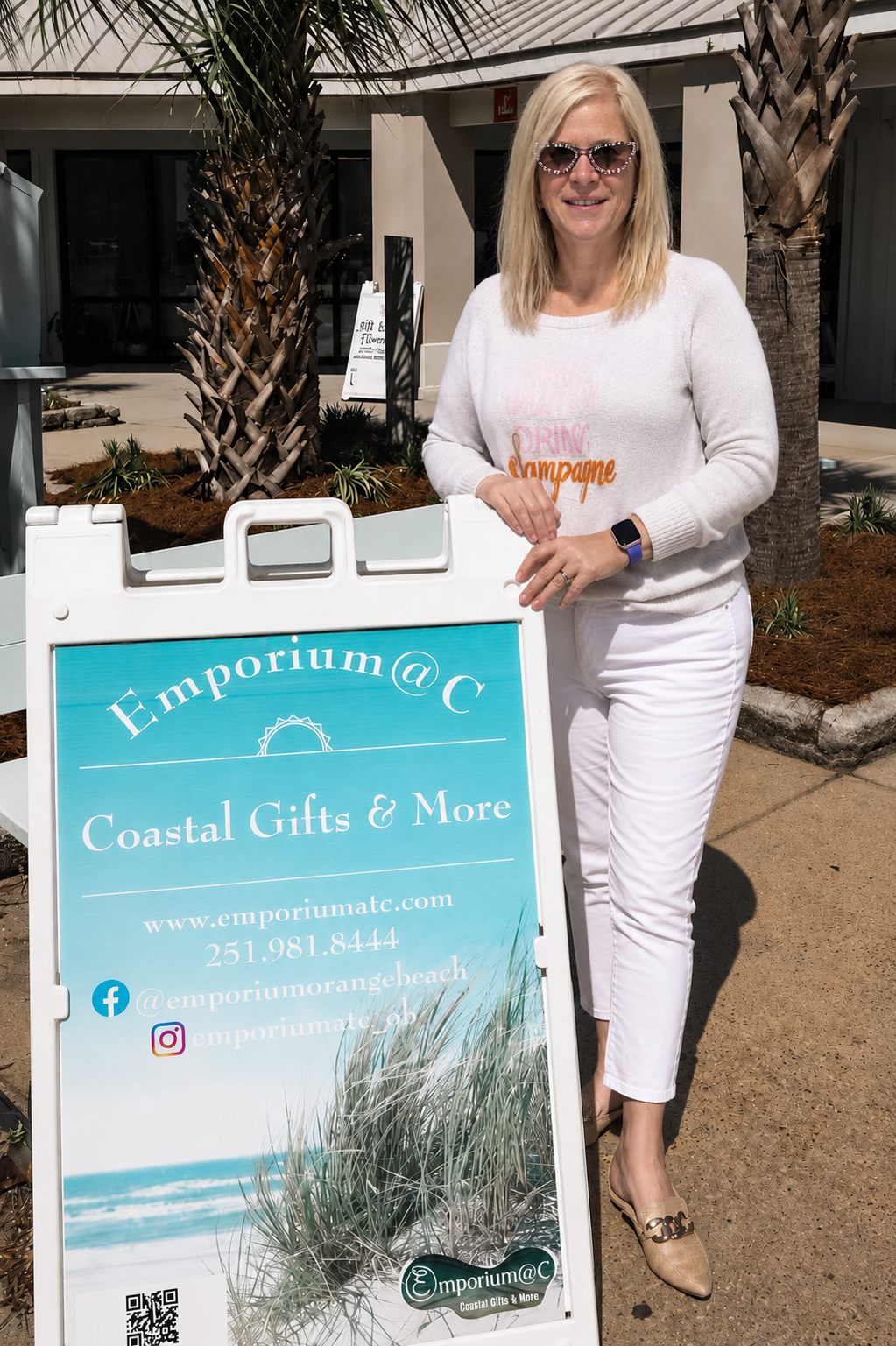 Woman owner standing next to a sign for Emporium @ C Coastal Gifts & More.