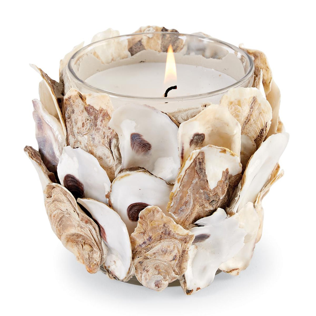 A lit white wax candle in a clear glass tumbler fully encrusted with real oyster shells in natural cream, tan, and brown tones.