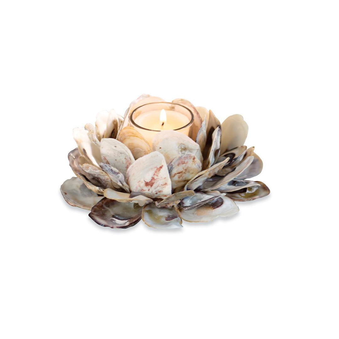 A flower-shaped candle holder made from layered natural oyster shells in cream, taupe, grey, and blush tones, holding a lit tealight candle in a clear glass votive cup.