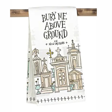A white kitchen towel hanging from a wooden rod, printed with the phrase 'BURY ME ABOVE GROUND IN NEW ORLEANS' and a line-art illustration of ornate New Orleans cemeteries in beige, black, and light blue.