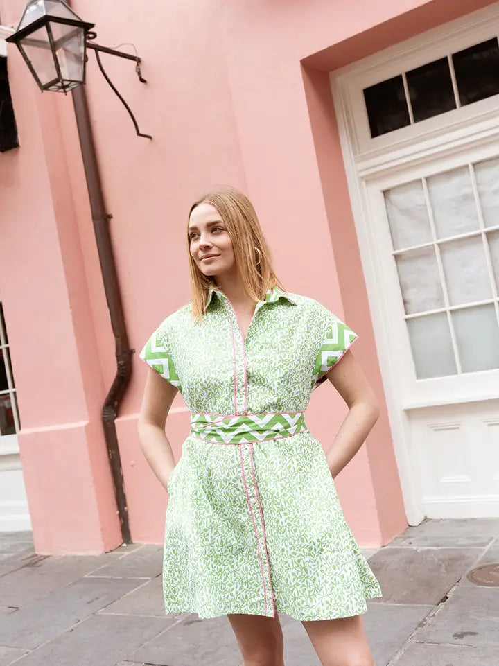 A woman wearing a short-sleeve A-line shirt dress in a green-and-white botanical print with chevron trim at the cap sleeves and waistband and pink piping along the front placket.