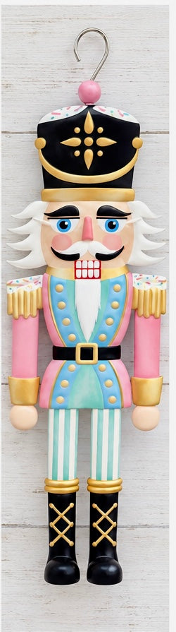 A flat, sign-style nutcracker soldier ornament in pastel pink, mint green, and gold, featuring a black crown with a gold star medallion, sprinkle-style trim on the hat and shoulders, gold buttons, a black belt with a gold buckle, striped trousers, and a metal hanging hook at the top.