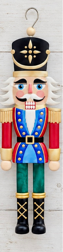 A flat, sign-style nutcracker soldier ornament in pastel pink, mint green, and gold, featuring a black crown with a gold star medallion, sprinkle-style trim on the hat and shoulders, gold buttons, a black belt with a gold buckle, striped trousers, and a metal hanging hook at the top.