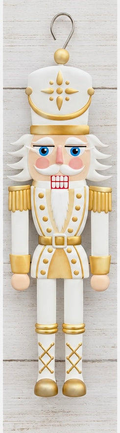 A flat, sign-style nutcracker soldier ornament in pastel pink, mint green, and gold, featuring a black crown with a gold star medallion, sprinkle-style trim on the hat and shoulders, gold buttons, a black belt with a gold buckle, striped trousers, and a metal hanging hook at the top.