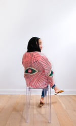 A woman seated on a clear chair facing away from the camera, wearing a cropped dusty pink velvet jacket with a large multicolor embroidered suzani-style medallion motif on the back.