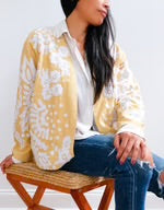 A woman seated on a wooden bench wearing a short wheat-yellow jacket with a large white floral suzani print, open collar, and button-front, styled over a white shirt and jeans.