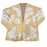A woman seated on a wooden bench wearing a short wheat-yellow jacket with a large white floral suzani print, open collar, and button-front, styled over a white shirt and jeans.