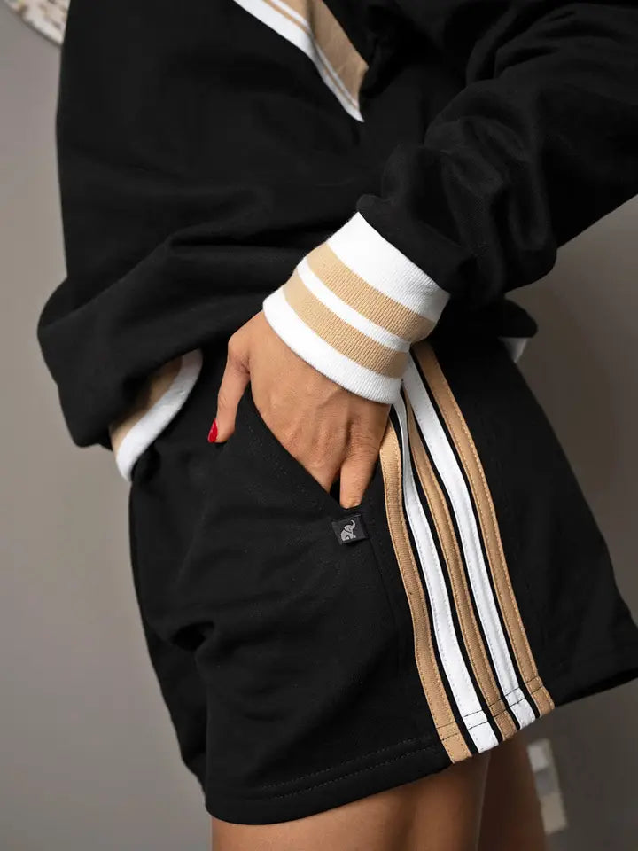 Black lounge shorts with tan and white vertical stripe tape along the outer side seam, a side hip pocket, and a small woven logo patch at the hip.