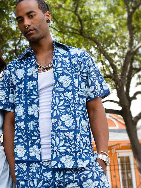 A man wearing a short-sleeve camp-collar button-up shirt in a bold blue and white botanical floral print, worn open over a white fitted tee.
